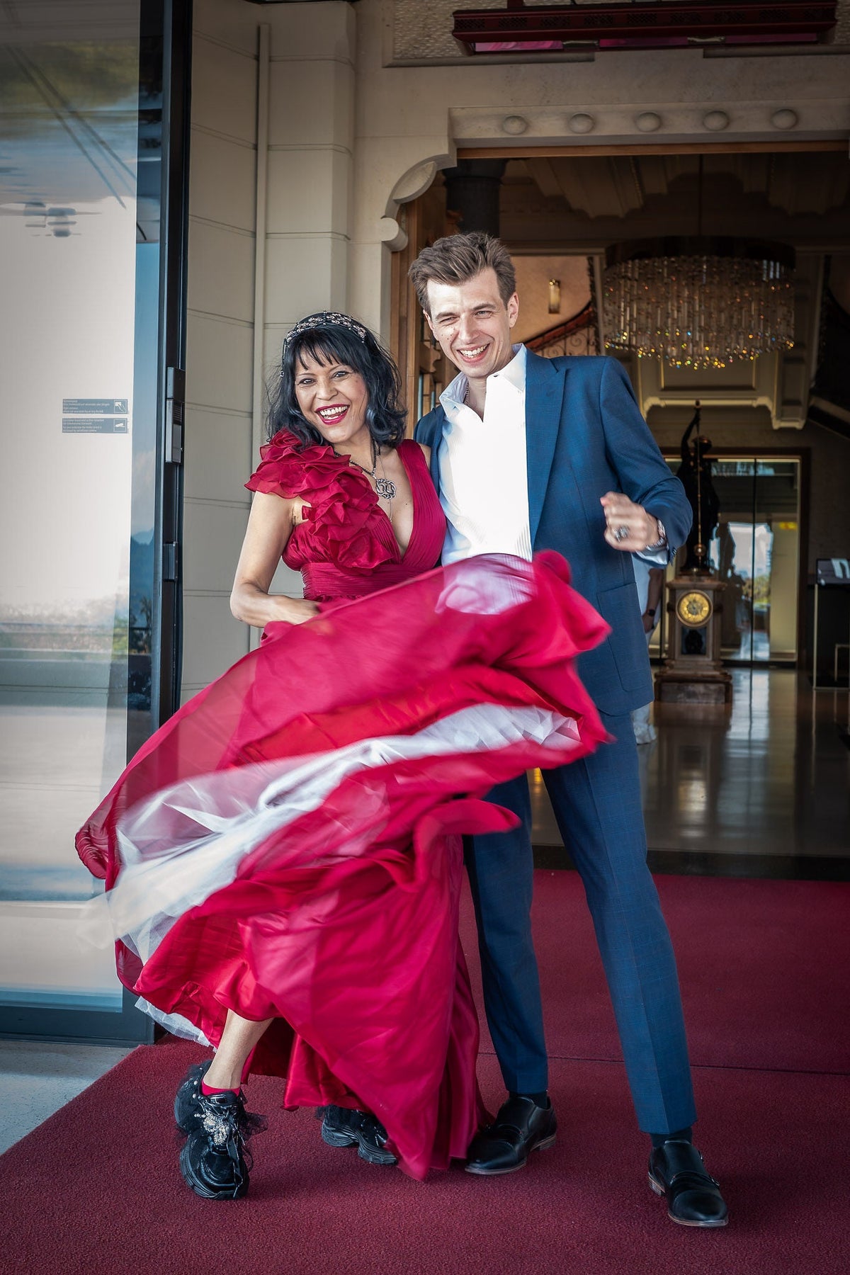 Natalie in a flowing red dress and Danny in a blue suit, smiling together on their wedding day.